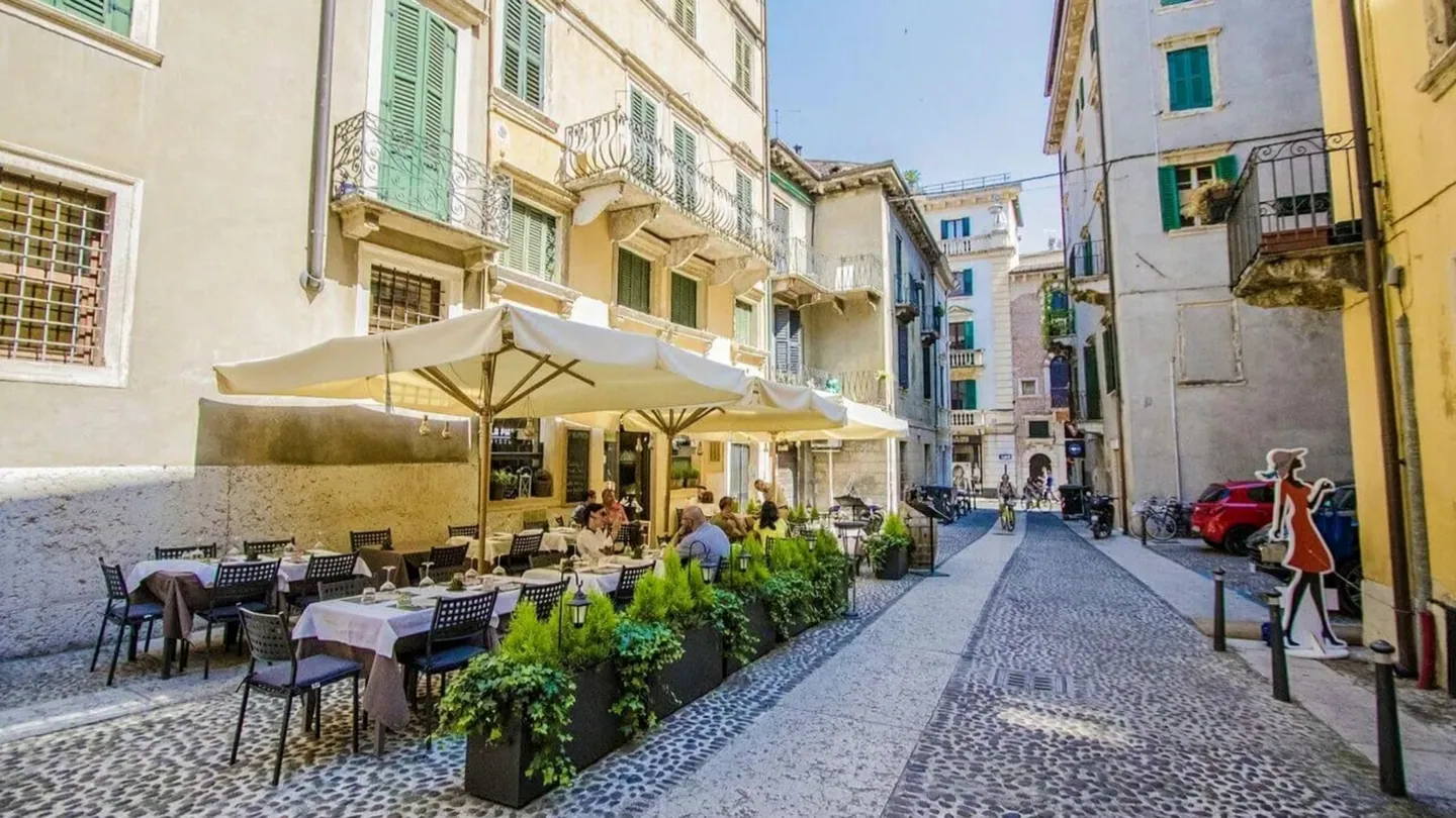 Il plateatico della Piazzetta in Corte San Giovanni in Foro, a Verona: tavoli apparecchiati sotto gli ombrelloni, piante verdi e le case con le persiane verdi